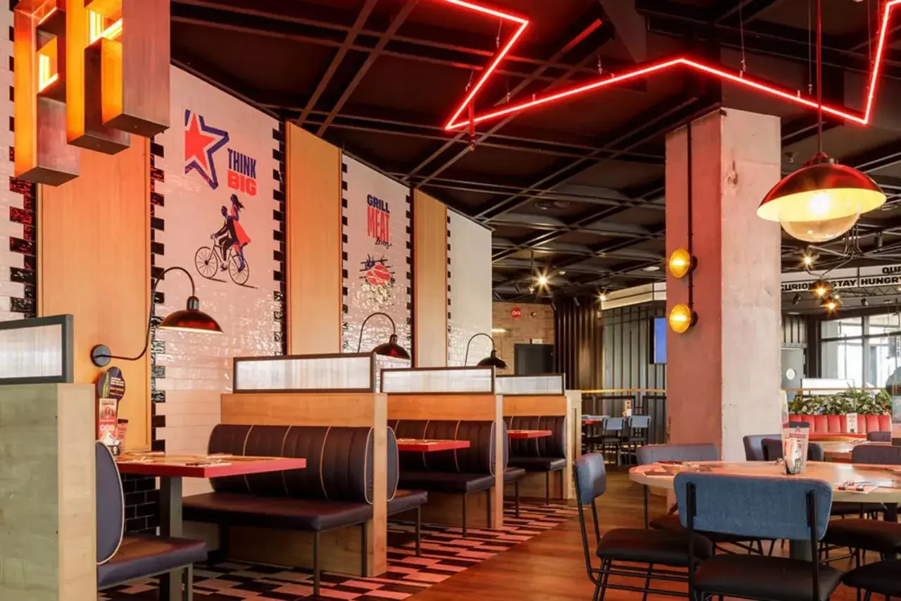 Interior de restaurante con cabinas, mesas y decoración moderna. Las paredes tienen murales inspiradores, como "Think Big". Las luces rojas de neón añaden un toque vibrante. Ideal para leer reseñas de Foster's Hollywood.