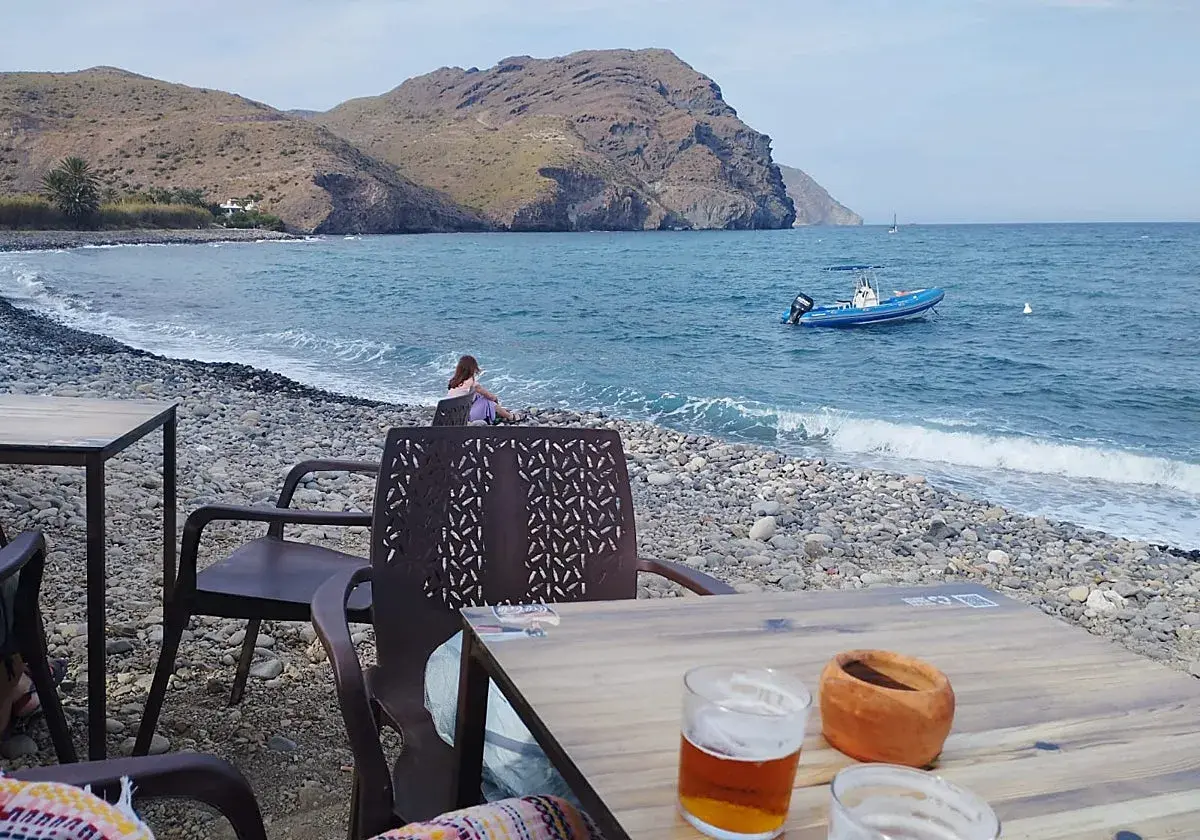 Una persona contempla el mar en la playa de las negras opiniones. Un bote azul descansa en el agua, mientras las olas acarician la costa pedregosa.
