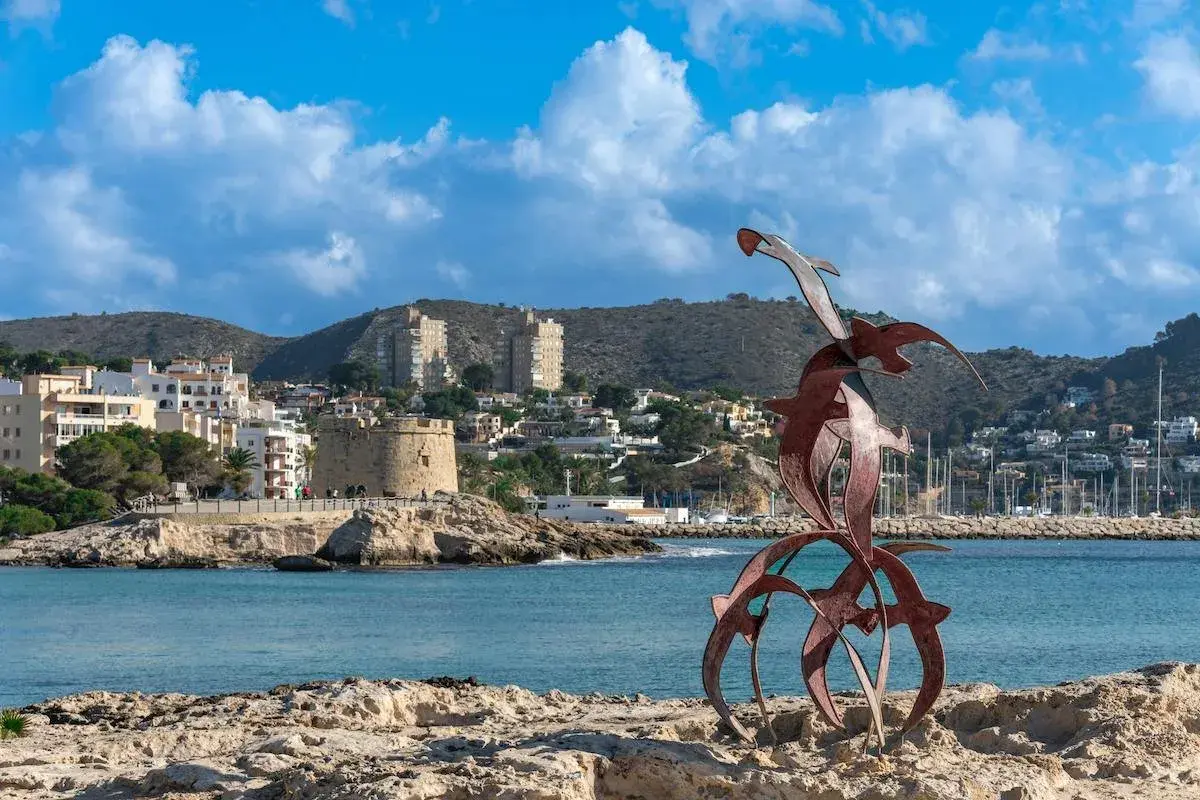 Escultura de aves sobre rocas, con el pueblo de Moraira y su torre defensiva al fondo.