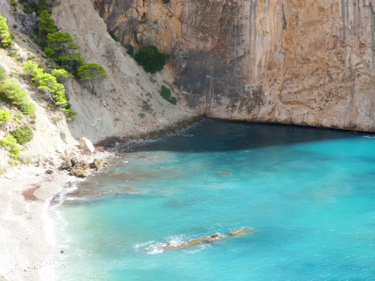 Aguas turquesas bañan la cala racó del corb, rodeada de imponentes acantilados y vegetación verde.