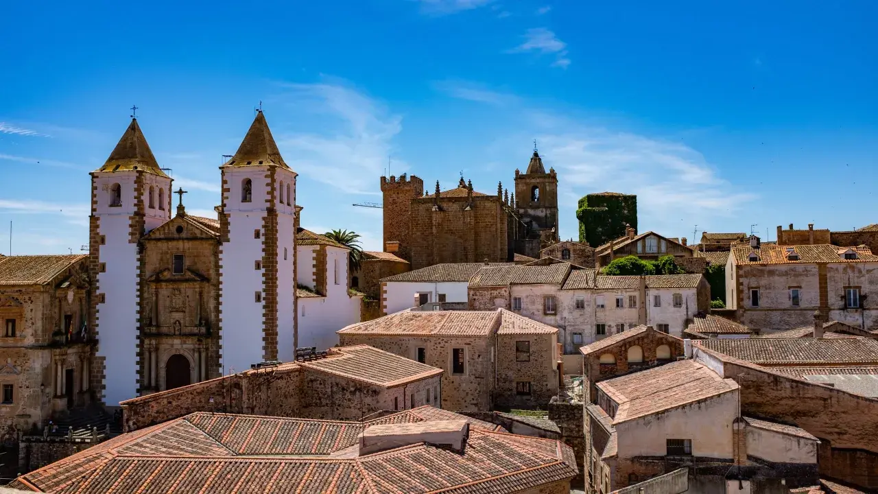 Vistas de tejados y edificios históricos, incluyendo iglesias con torres blancas y campanarios, bajo un cielo azul. Un lugar ideal que ver en Cáceres provincia.