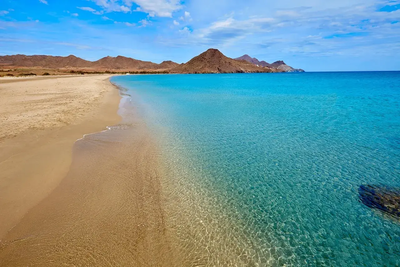Arena dorada y aguas cristalinas en una de las playas bonitas en España, con montañas áridas al fondo bajo un cielo azul.