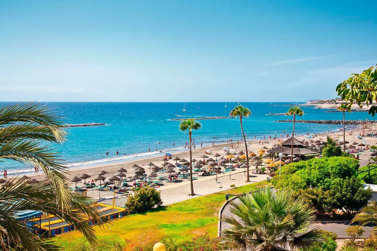 Un día soleado en el paseo marítimo de Playa de las Américas, con gente disfrutando de la playa, sombrillas y palmeras.