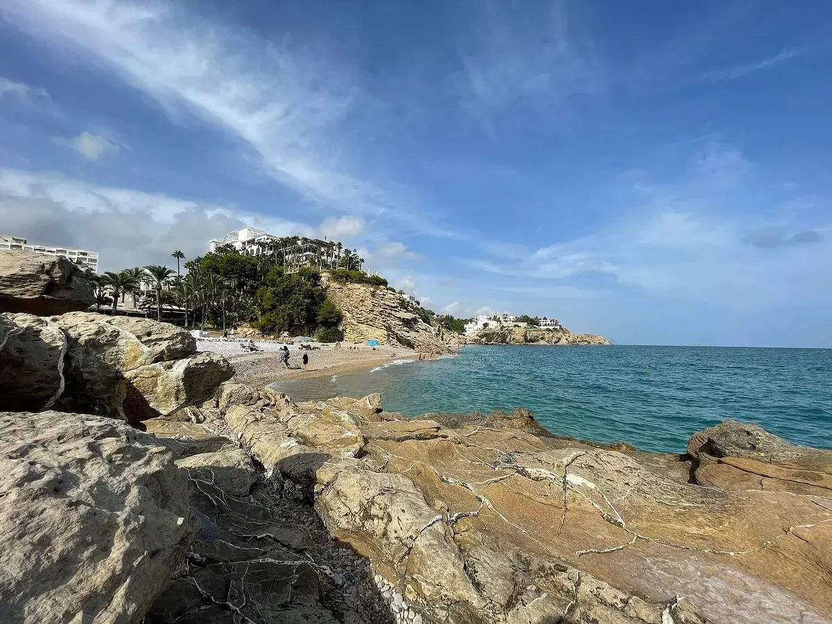 Rocas doradas en primer plano, la caleta Villajoyosa con gente disfrutando del mar azul y edificios blancos en la colina.