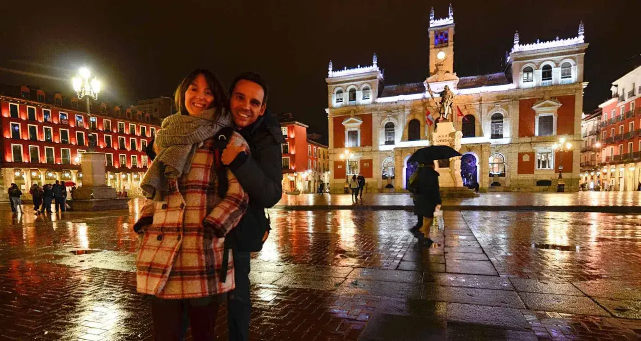 Pareja sonriente en la Plaza Mayor de Valladolid, un lugar bonito que ver en Valladolid, con edificios iluminados y el suelo mojado por la lluvia.