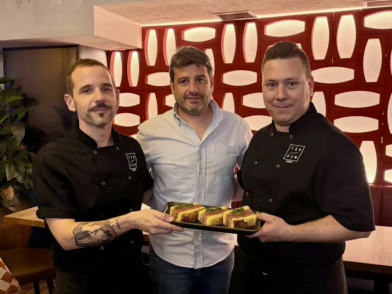 Tres hombres posan con un plato de aperitivos en un mesón. Las fotos capturan el momento.