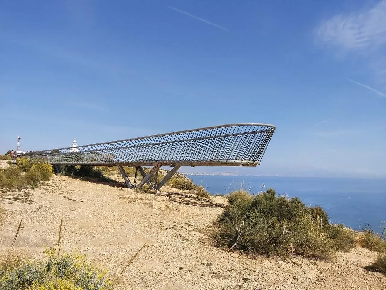 El mirador del faro de Santa Pola se proyecta sobre el mar azul, con vegetación árida y un cielo despejado.