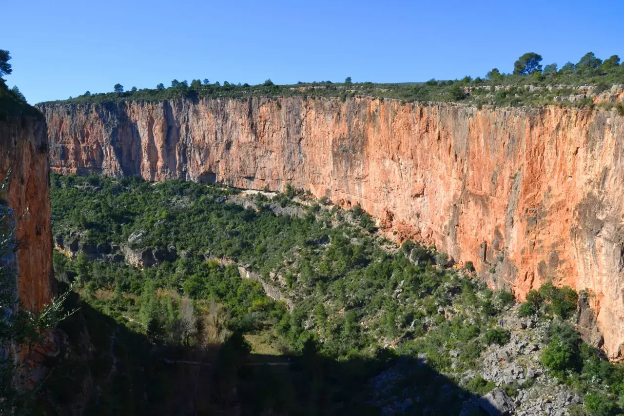 Impresionante pared rocosa anaranjada en la ruta de los calderones Chulilla, con vegetación verde cubriendo el fondo del cañón.