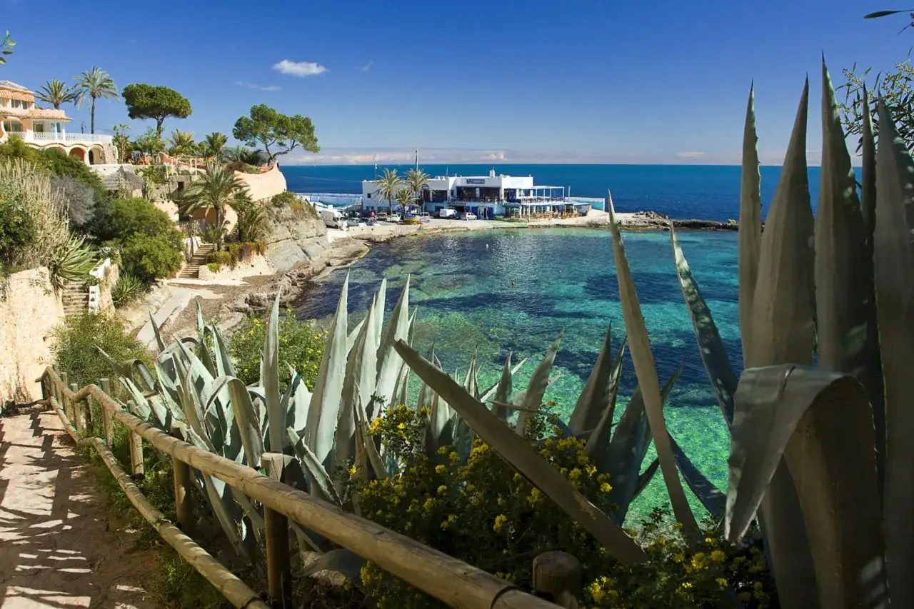 Un camino con barandilla de madera y agaves conduce a una cala con aguas turquesas. Un restaurante blanco se asoma al mar. &iexcl;Descubre qu&eacute; ver en Benissa!
