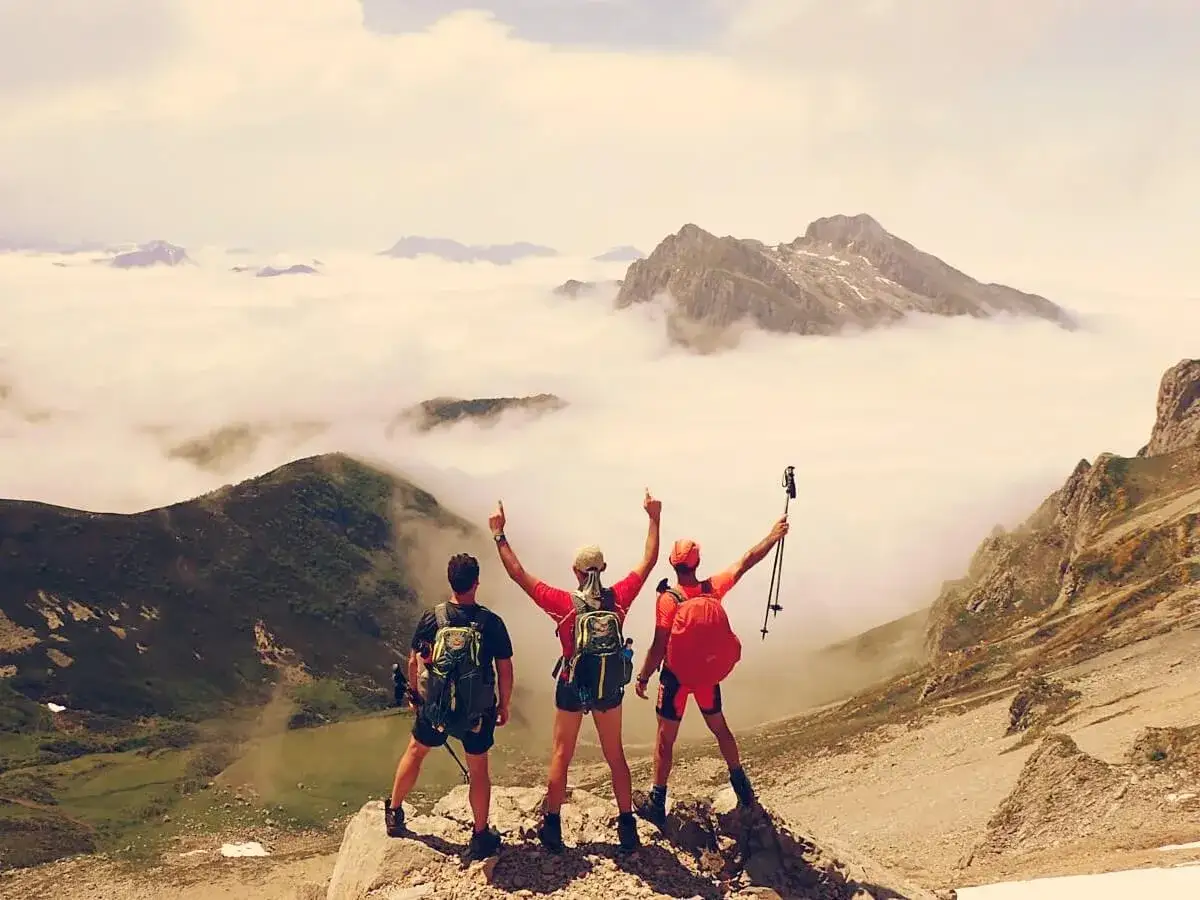 Tres excursionistas celebran en la cima, con los Picos de Europa cubiertos de nubes. ¡Una aventura inolvidable!