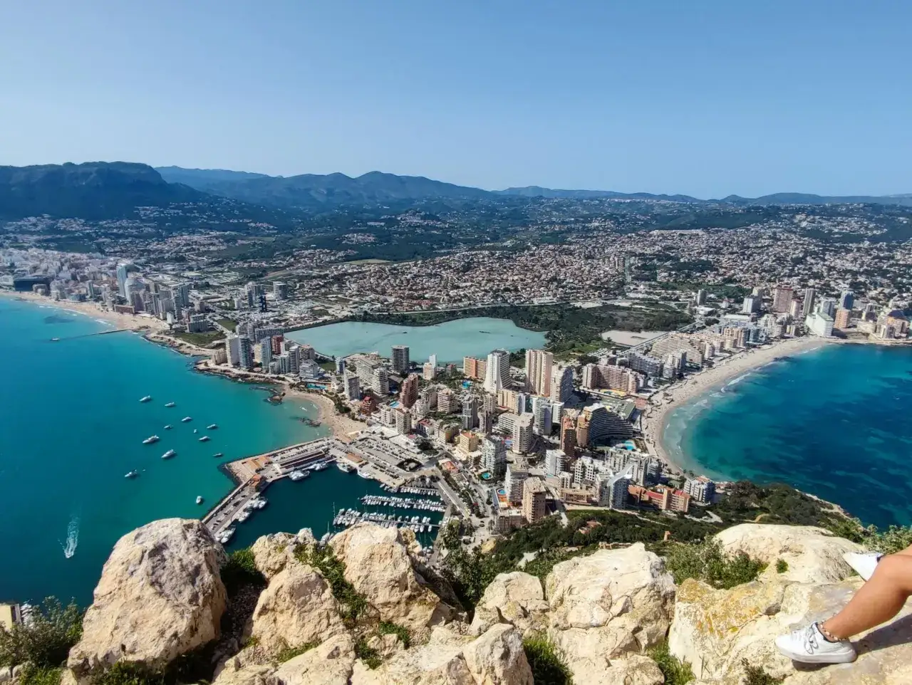 Vista a&eacute;rea de la cala del penyal, con el mar turquesa, el puerto deportivo, la playa y la ciudad extendi&eacute;ndose hasta las monta&ntilde;as.
