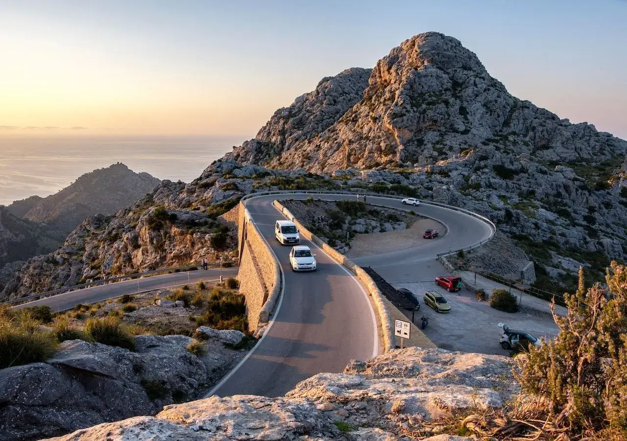 Curva sinuosa en el mirador Sa Foradada, con coches circulando y el mar al fondo al atardecer.