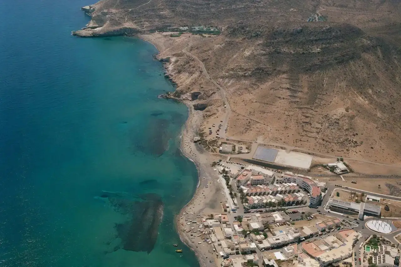 Vista aérea de la playa de las negras opiniones, con aguas turquesas, arena, rocas y un pueblo costero.