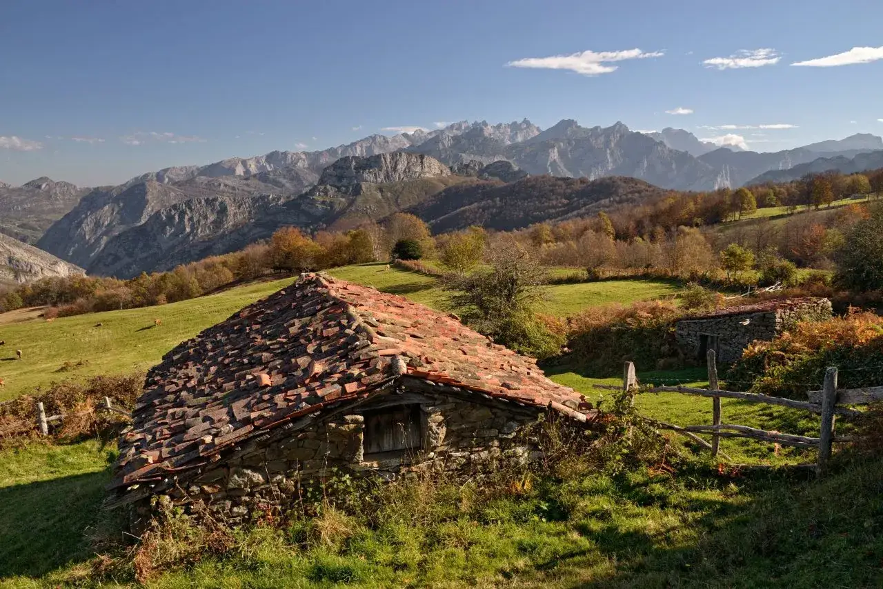 Majestuosas montañas en el parque natural de Ponga. Un antiguo refugio de piedra con tejado de tejas rojas se alza en un prado verde, con otras construcciones al fondo.