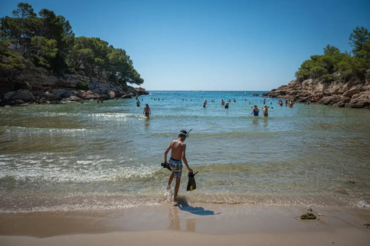 Un niño con equipo de snorkel se adentra en el mar en el paseo marítimo de l'Ametlla de Mar. Gente disfrutando del agua en un día soleado.