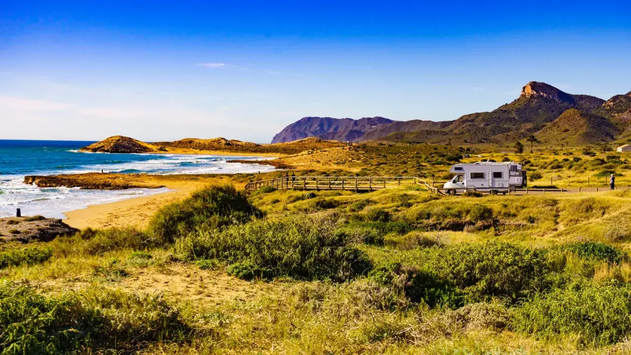 Autocaravana en la costa, con el mar azul y el monte de las cenizas al fondo.