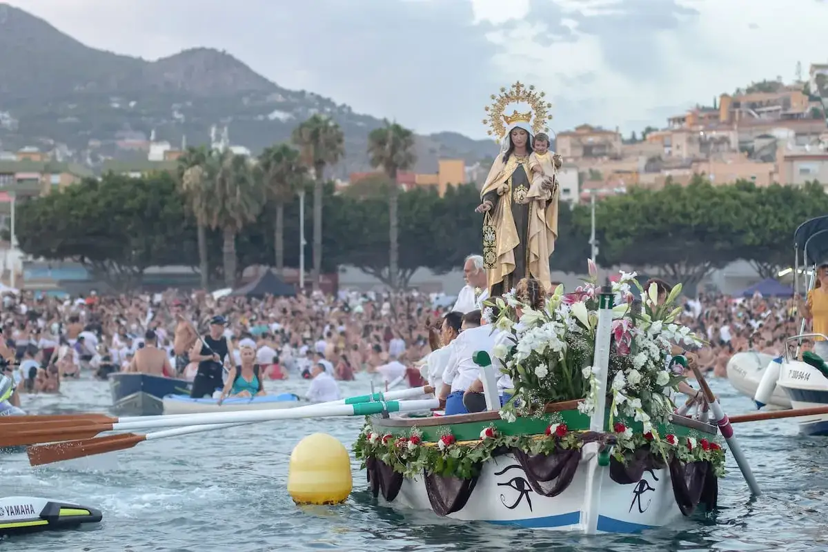 Procesión marítima de la Virgen del Carmen. Una multitud celebra las fiestas del Carmen en el mar, rodeando una barca adornada con flores.