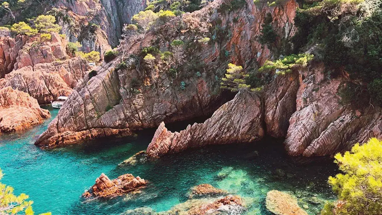 Aguas turquesas bañan las rocas escarpadas de la Costa Brava, España. Un pequeño barco navega en una cala escondida entre acantilados cubiertos de vegetación.