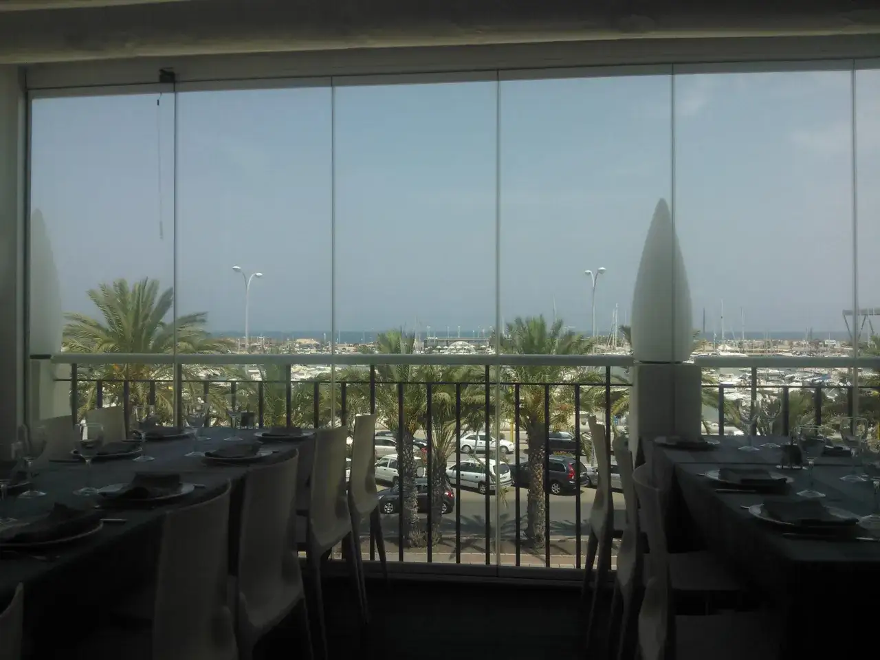 Mesas puestas con vistas al puerto deportivo de la barqueta Denia. Palmeras y barcos crean un ambiente vacacional.