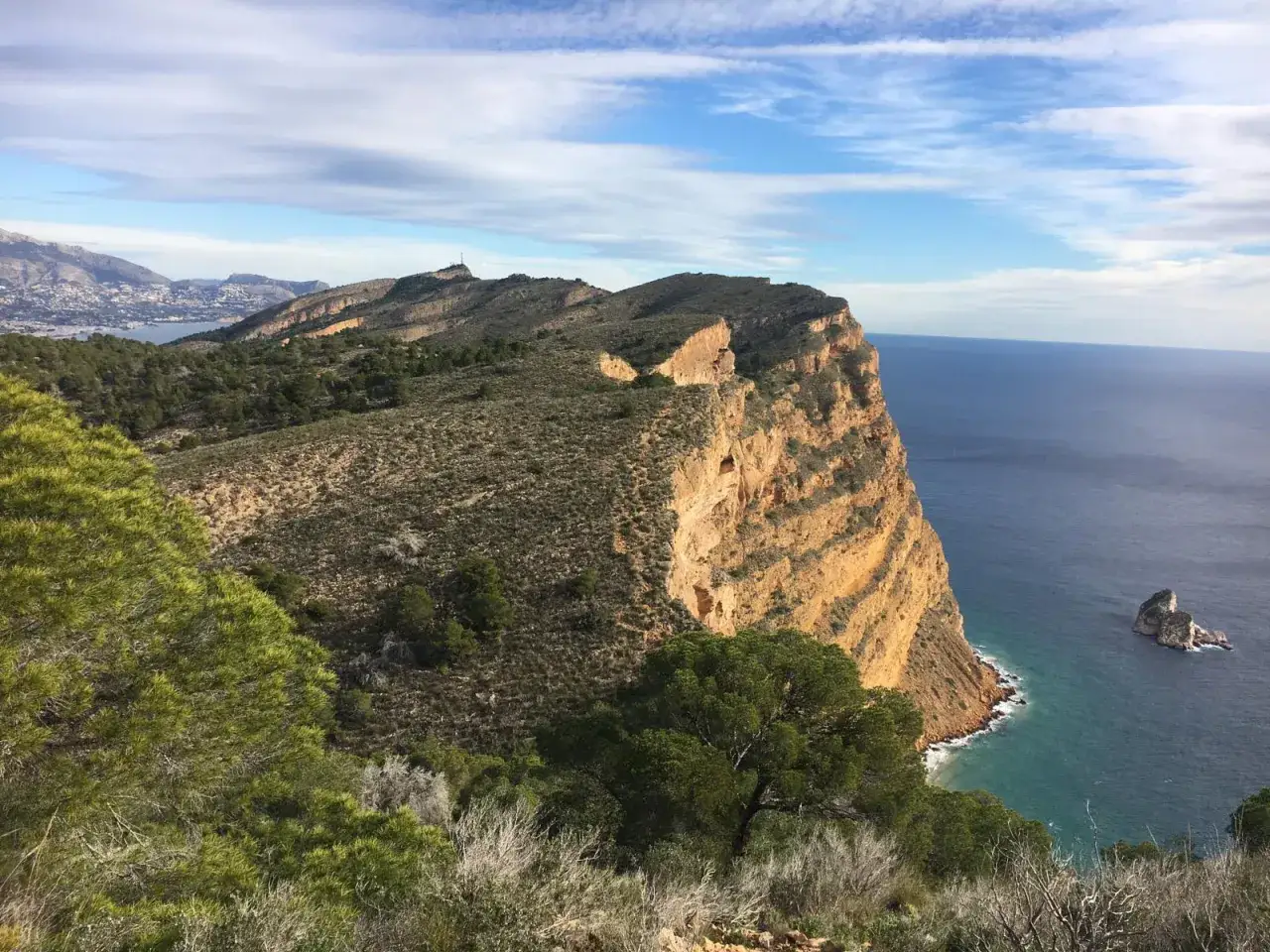 Vista de acantilados dorados y el mar azul, con vegetación mediterránea y la silueta de la cruz de Benidorm en la distancia.