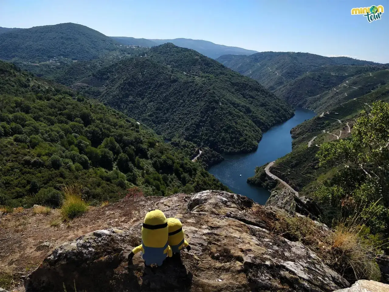 Dos minions contemplan el impresionante paisaje desde el mirador de Vilouxe, con un río serpenteando entre montañas verdes.