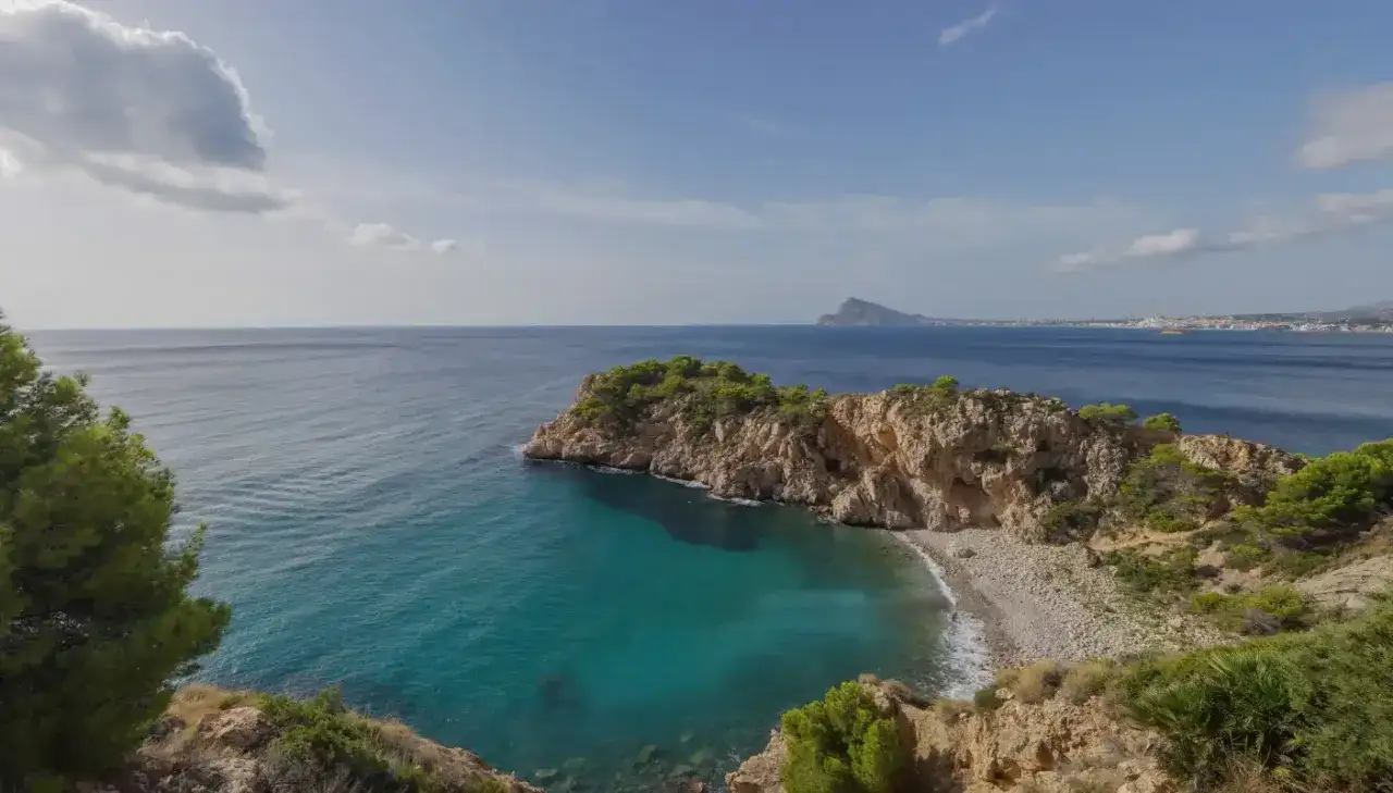 Vista de una cala con aguas turquesas y rocas cubiertas de vegetaci&oacute;n. Al fondo, el mar azul se extiende hasta la costa con edificios.