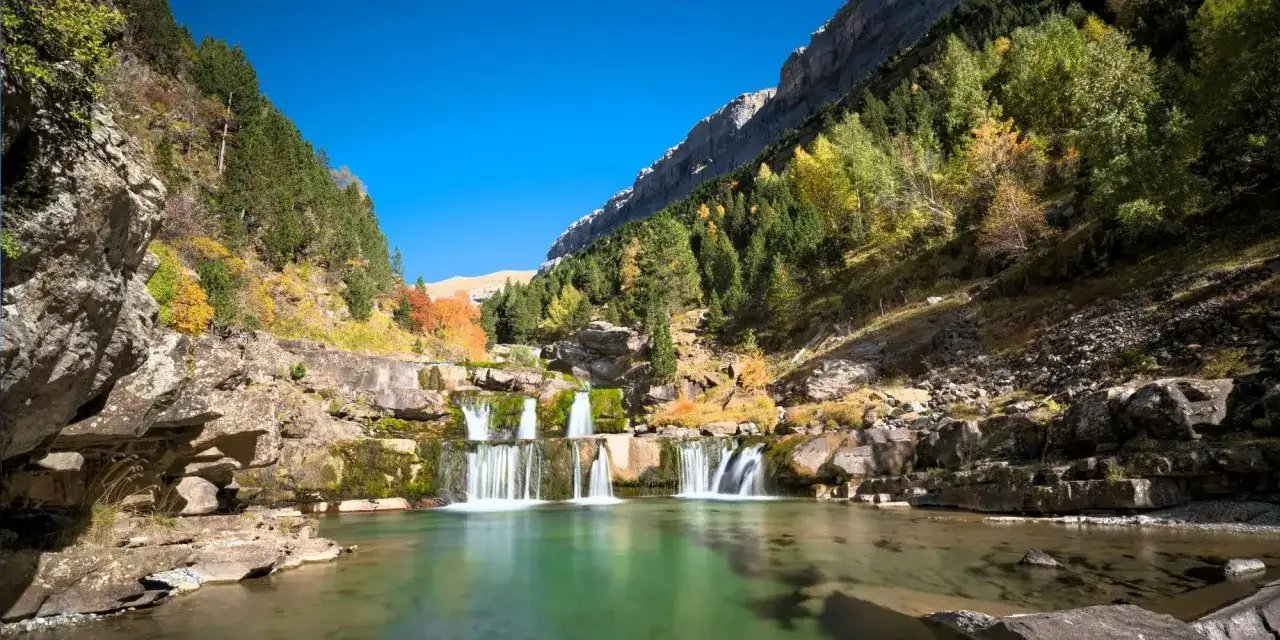 Cascada en un valle montañoso, ideal para rutas senderismo España. El agua verde turquesa fluye entre rocas y árboles otoñales.