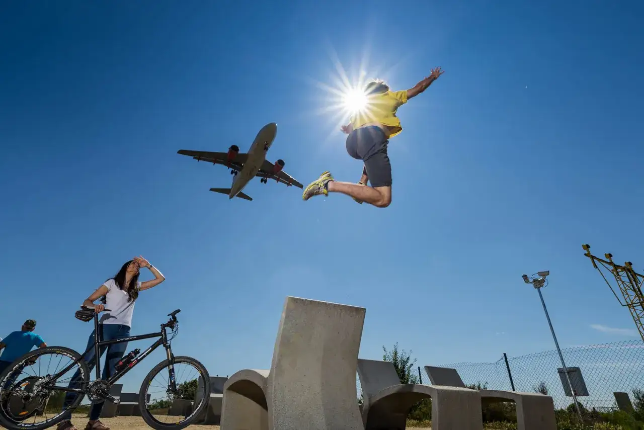 Un hombre salta de alegría bajo el sol mientras un avión pasa sobre el mirador de aviones de El Prat. Una mujer con bicicleta observa.