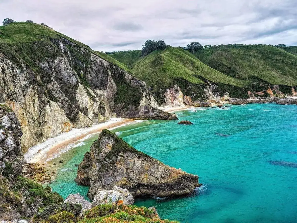 Acantilados verdes y rocosos bordean una playa de arena blanca y aguas turquesas, un paraíso entre las playas más bonitas de Asturias.