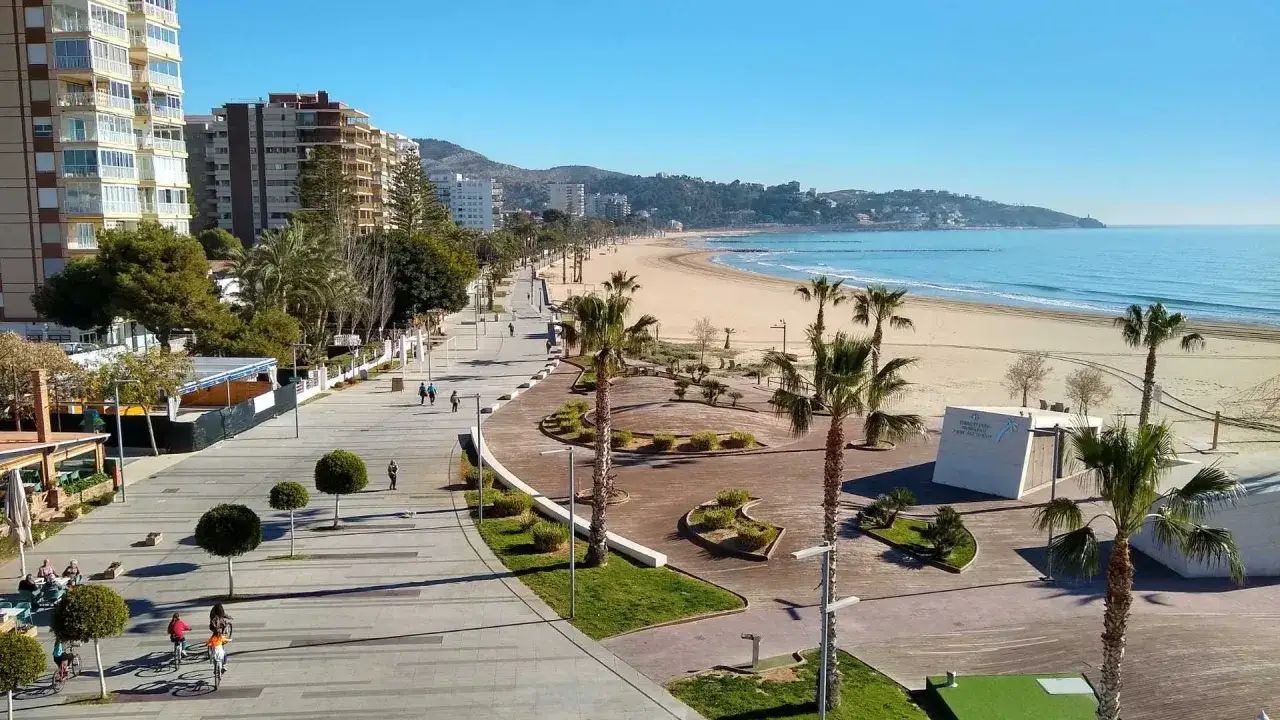 El benicàssim paseo marítimo, con palmeras y edificios altos, se extiende hacia una playa dorada y el mar azul bajo un cielo despejado.