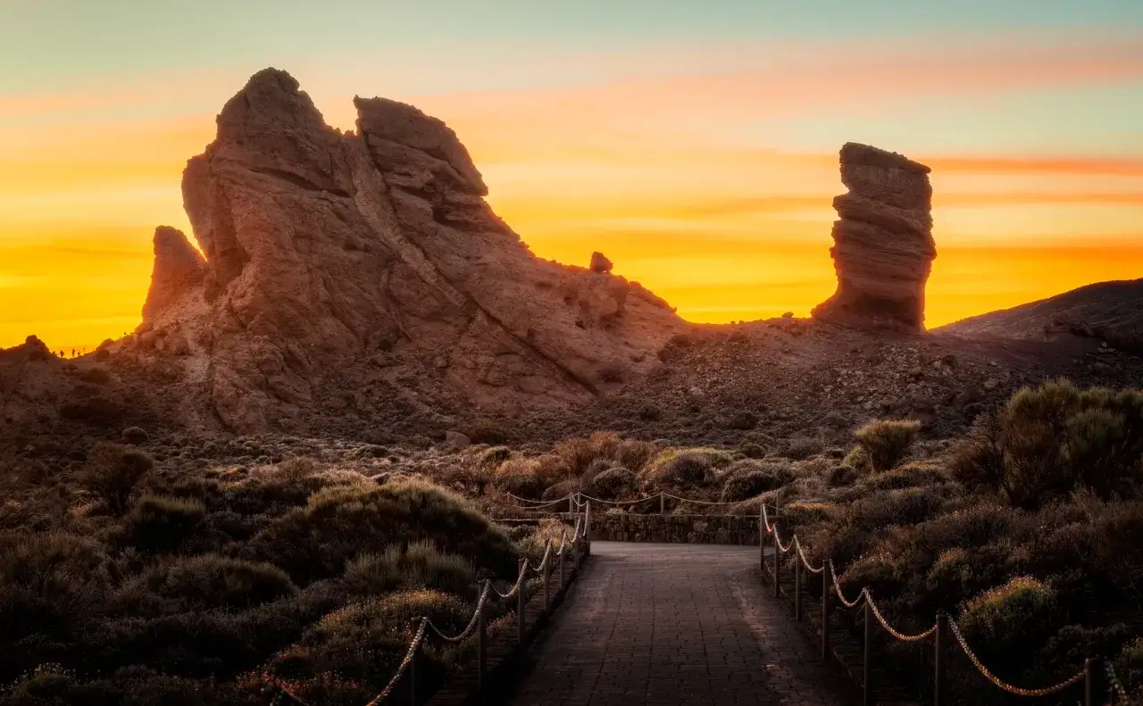 Camino de piedra hacia el Roque Cinchado al atardecer. El sendero serpentea entre matorrales bajo un cielo anaranjado.