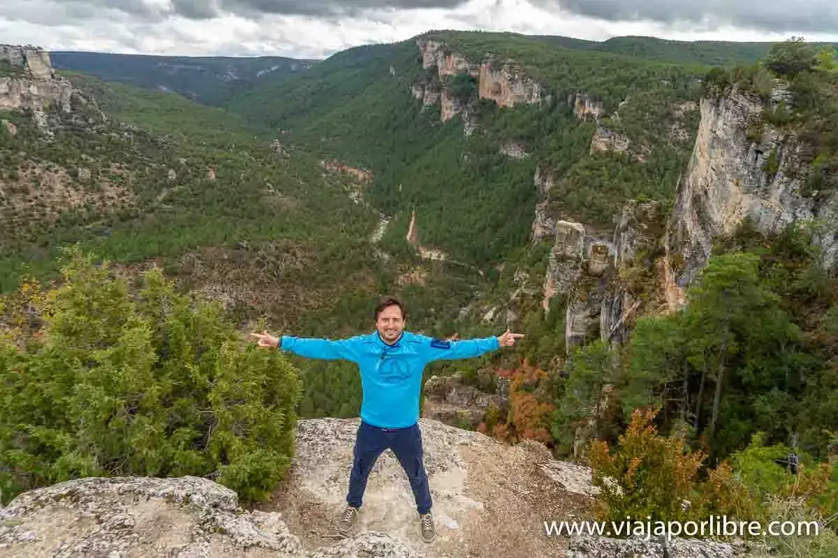 Hombre sonriente en un acantilado con un paisaje de cañón verde y rocoso. ¡Qué alto tajo que ver!