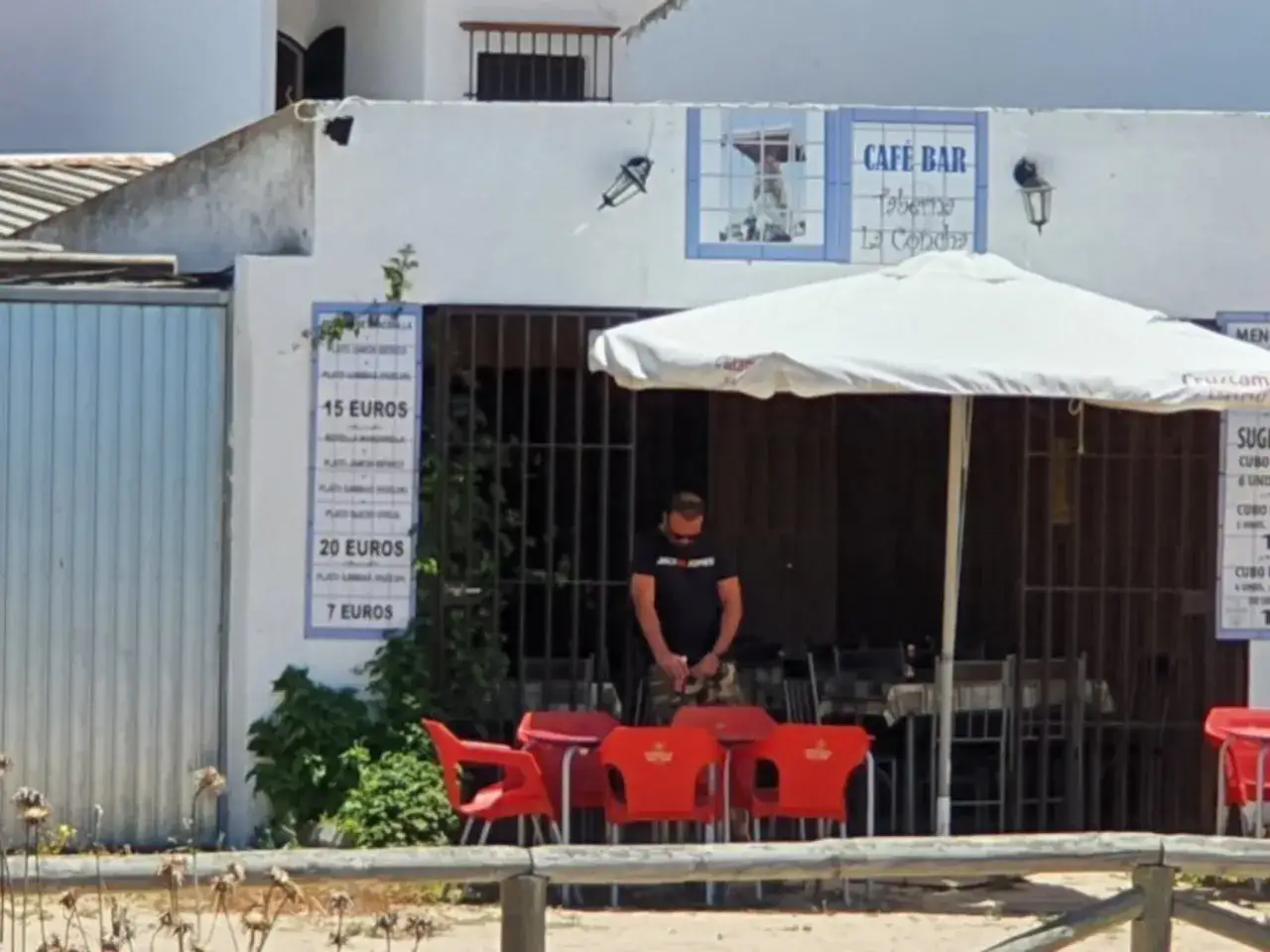 Un hombre mira su m&oacute;vil frente a la Taberna La Concha, con sillas rojas y un men&uacute; de precios visibles.