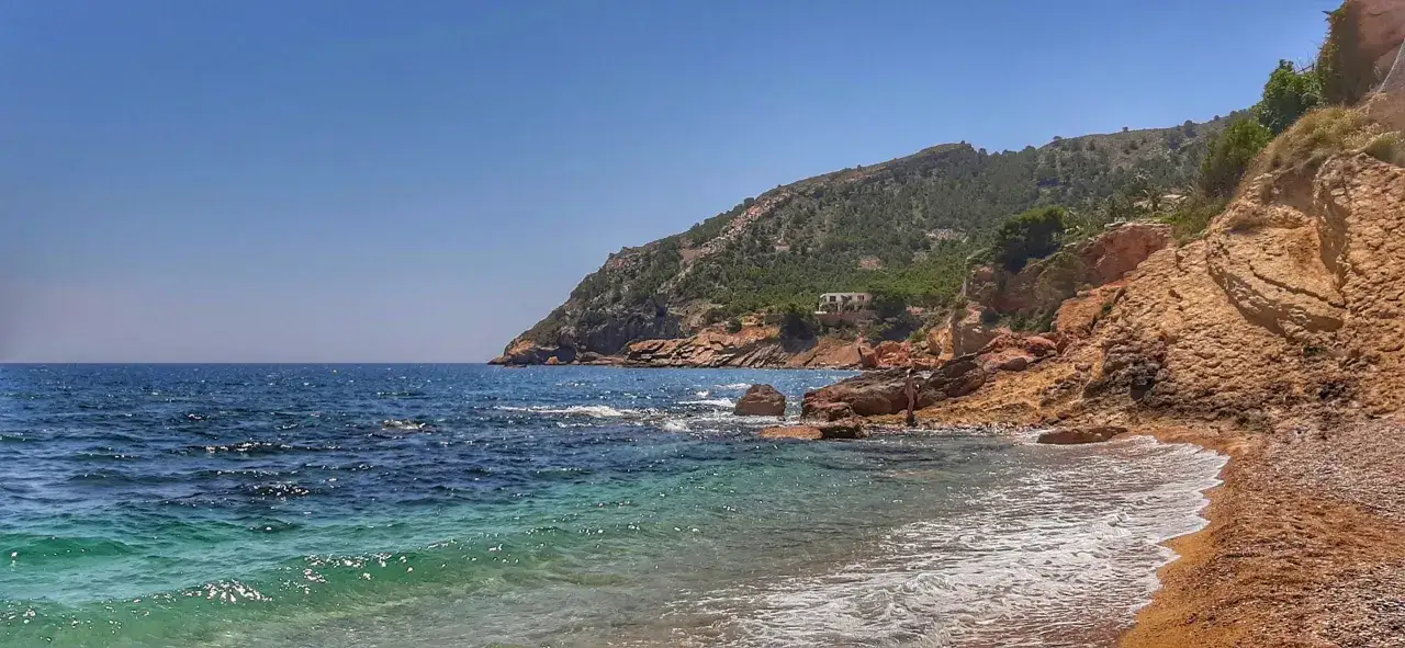 Aguas turquesas bañan la cala del metge, con rocas rojizas y una ladera verde coronando el paisaje.
