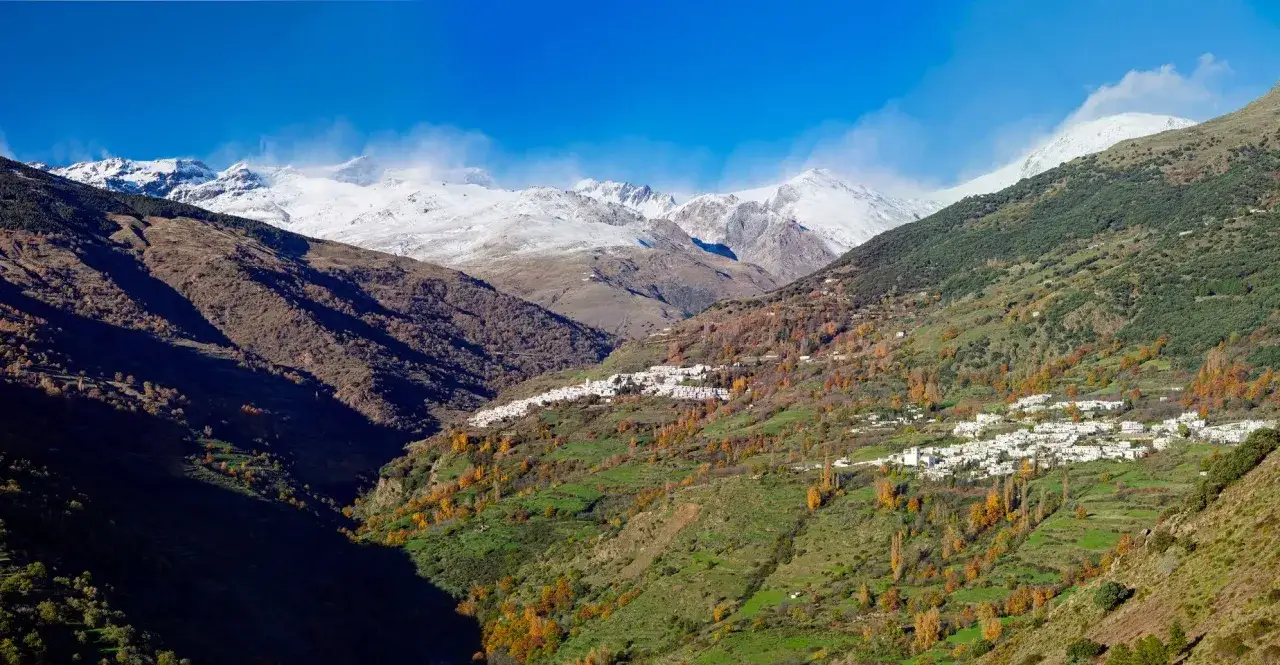 Pueblo blanco en ladera verde con montañas nevadas al fondo. Un destino encantador que ver cerca de Granada.