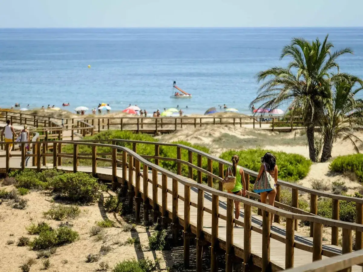 Dos mujeres caminan por un sendero de madera hacia la playa Los Arenales, con el mar azul y gente disfrutando del sol.