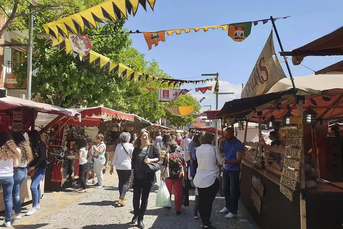 Gente pasea por un animado mercadillo medieval el &aacute;lamo, con puestos de artesan&iacute;a y comida bajo banderas coloridas.