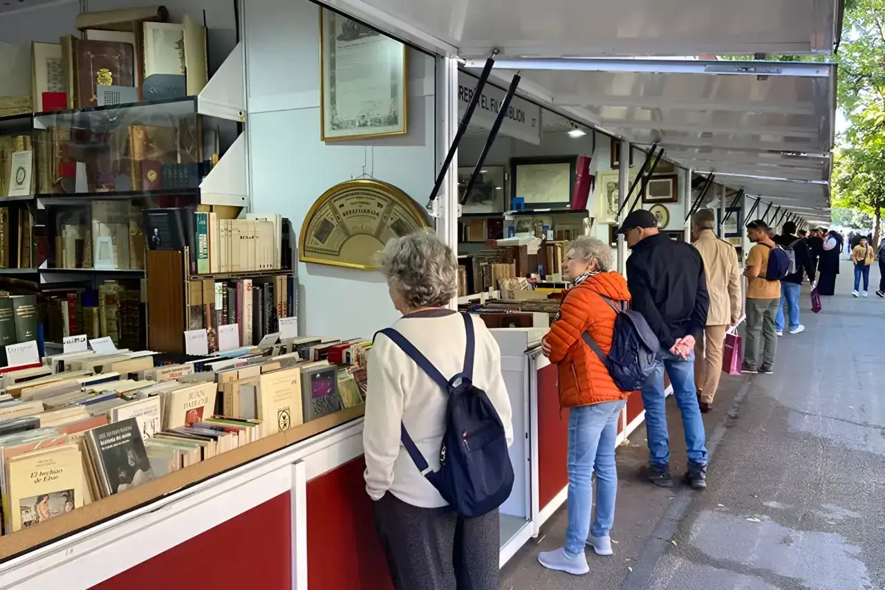 Gente buscando tesoros literarios en la feria del libro antiguo de Madrid. Estanterías repletas de tomos esperan ser descubiertos.