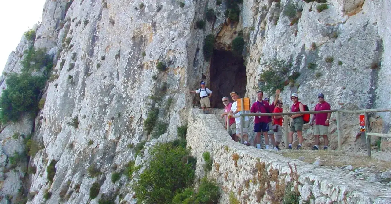 Grupo de excursionistas en el sendero del Peñón de Ifach, con una cueva en la roca y un muro de piedra.