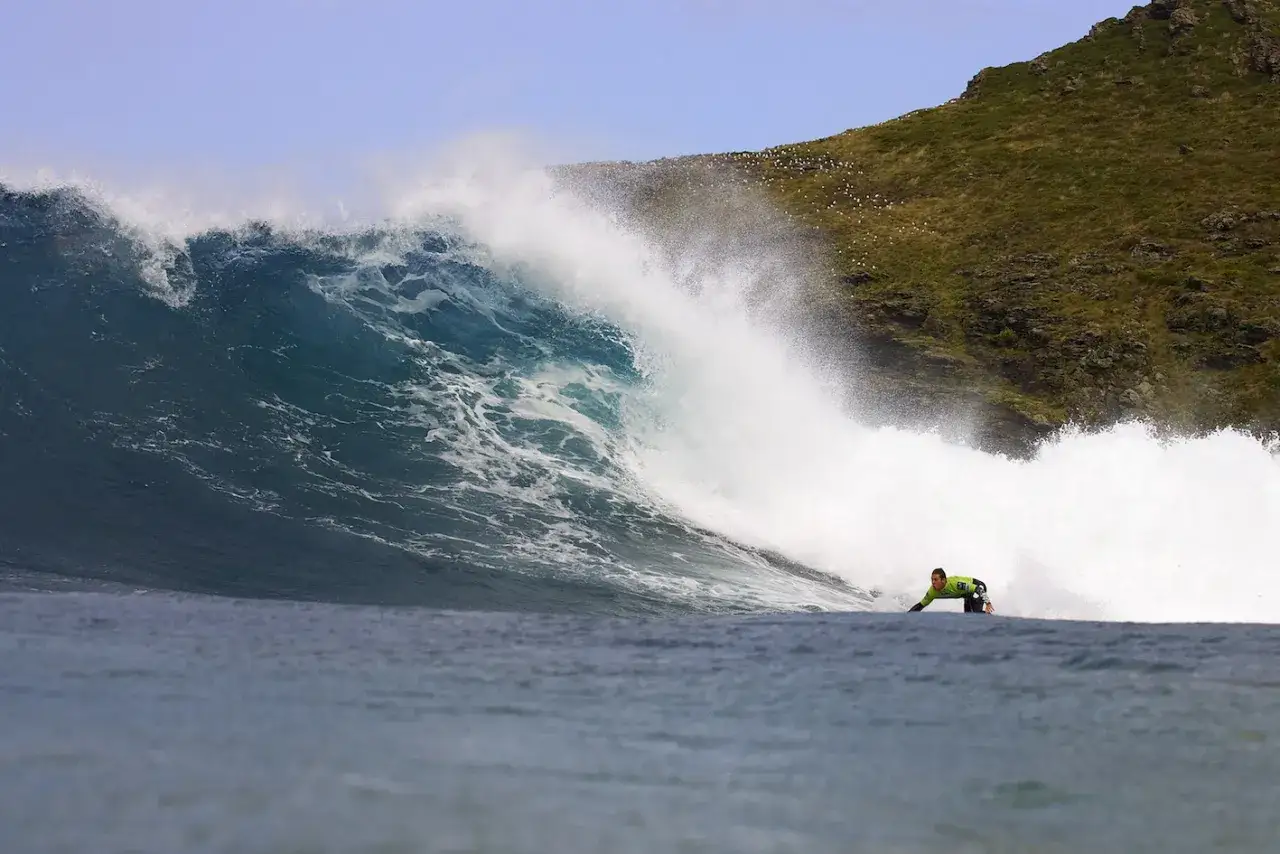 Surfista desafía una ola gigante en la playa de Pantín, con la costa verde y rocosa al fondo.