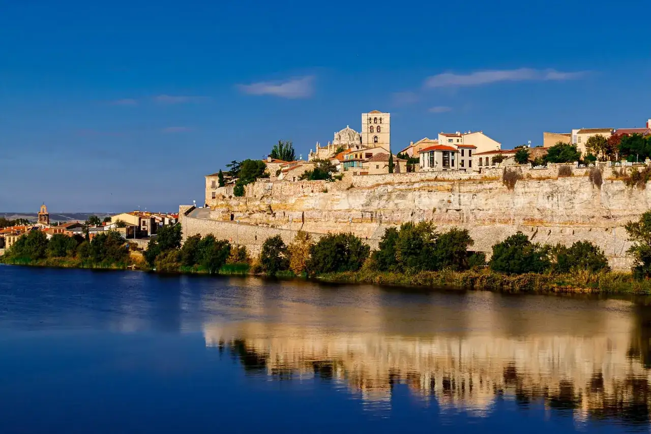 Impresionante vista de Zamora que ver: edificios históricos sobre un acantilado, reflejados en el río Duero bajo un cielo azul.