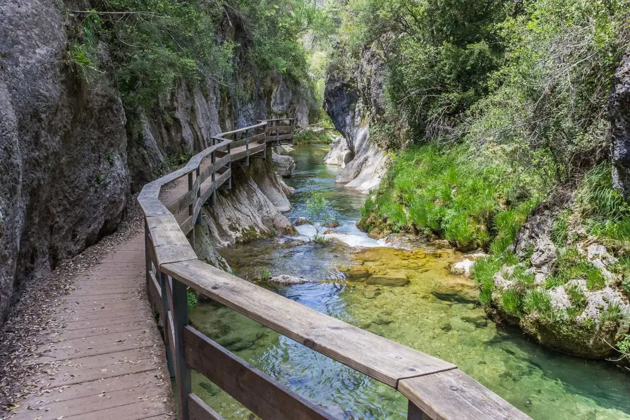 Sendero de madera serpentea junto a un río cristalino entre rocas y vegetación exuberante, un rincón idílico de los parques naturales de España.