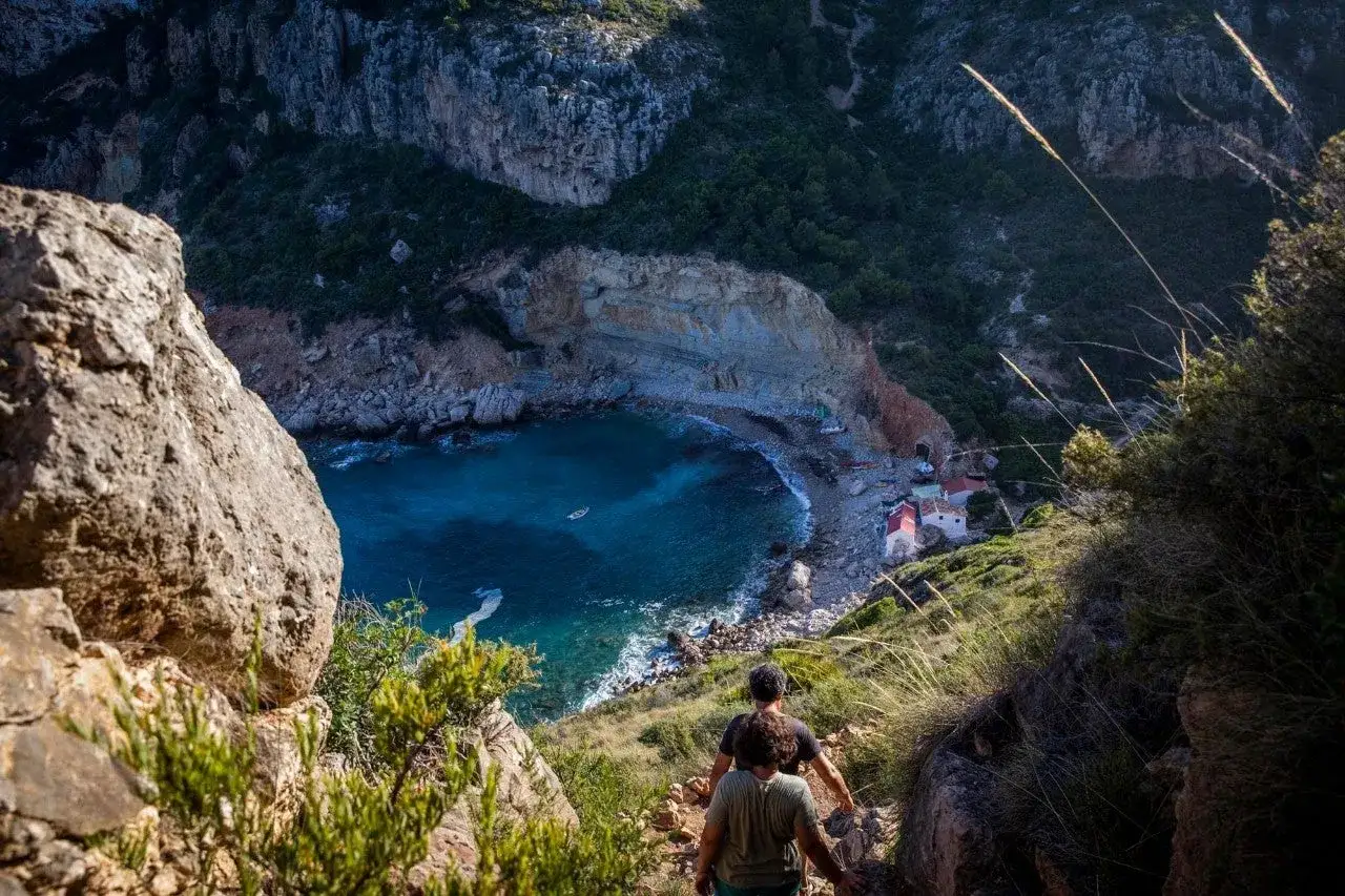 Dos excursionistas descienden por un sendero rocoso hacia la cala dels testos, una bah&iacute;a de aguas turquesas con casitas blancas y un peque&ntilde;o bote.