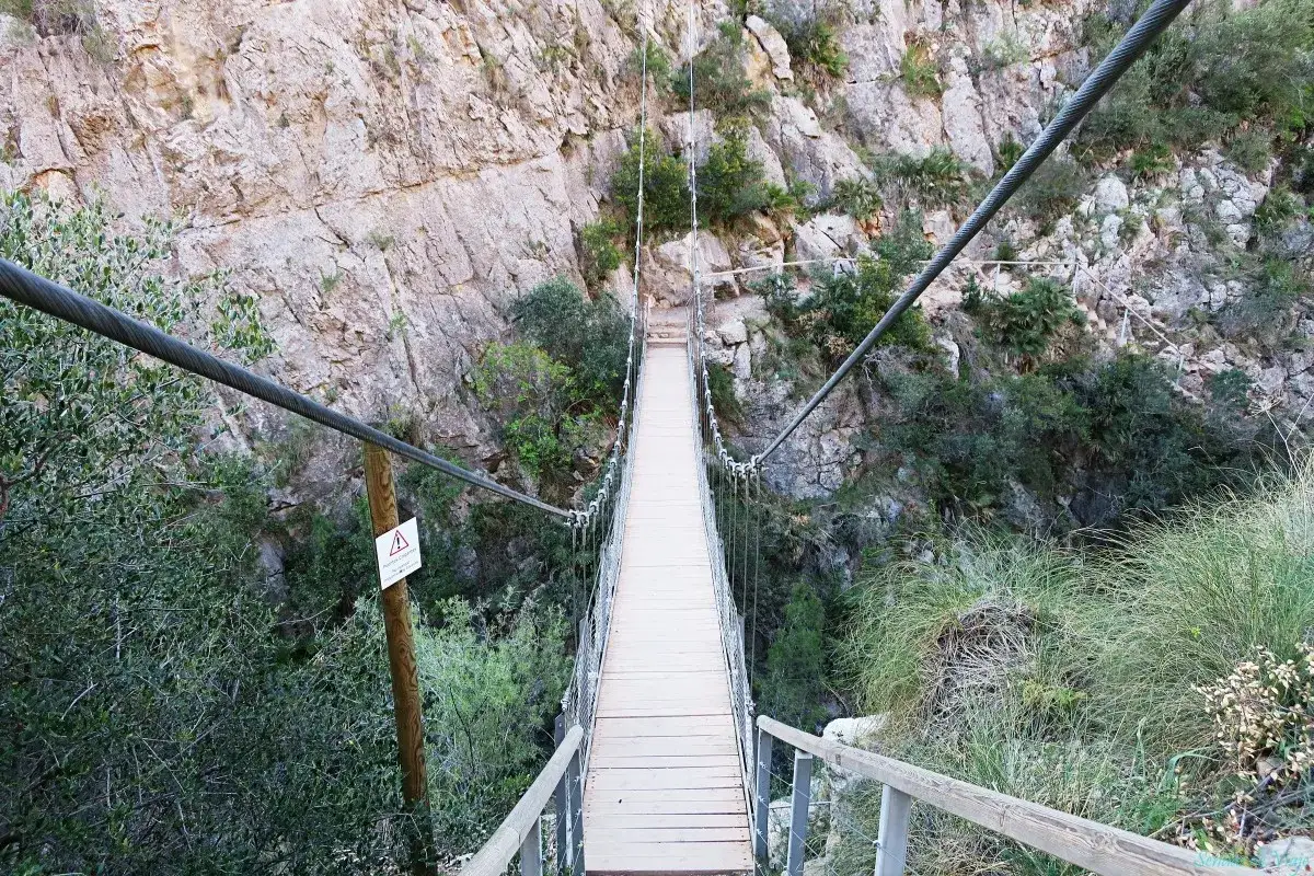 Puente colgante en la ruta de los calderones Chulilla, rodeado de vegetaci&oacute;n y acantilados rocosos.