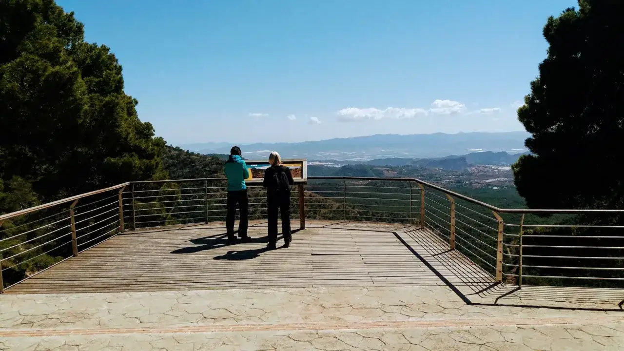 Dos personas observan un panel informativo en un mirador con vistas al estrecho de la Arboleja y un valle verde.