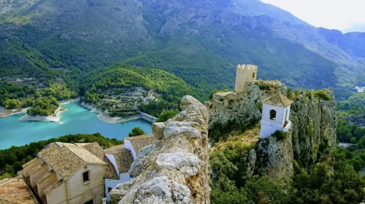Vista panorámica del embalse de Guadalest con su agua turquesa, rodeado de montañas verdes y el castillo en la cima.