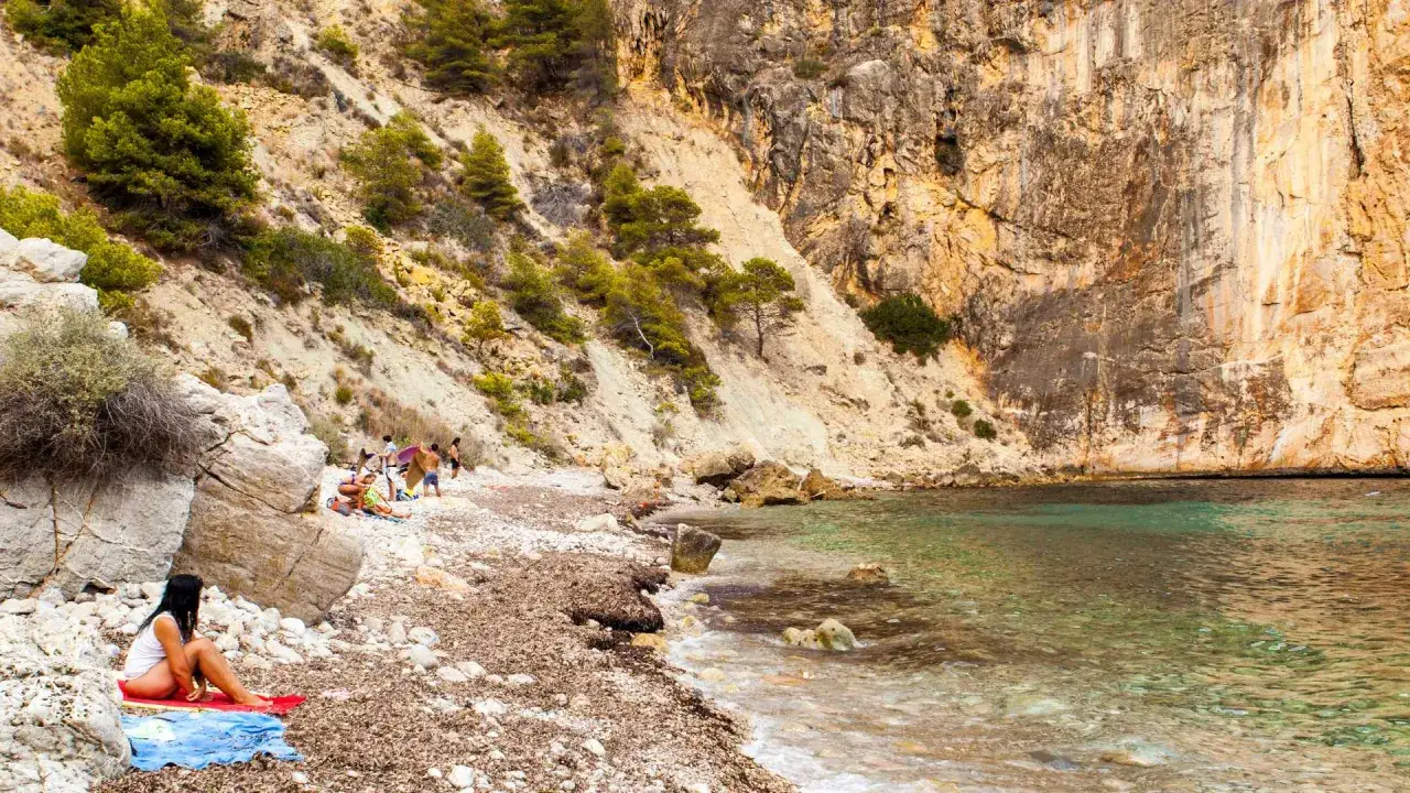 Una mujer descansa en la playa de cala raco del corb, con acantilados rocosos y aguas cristalinas.