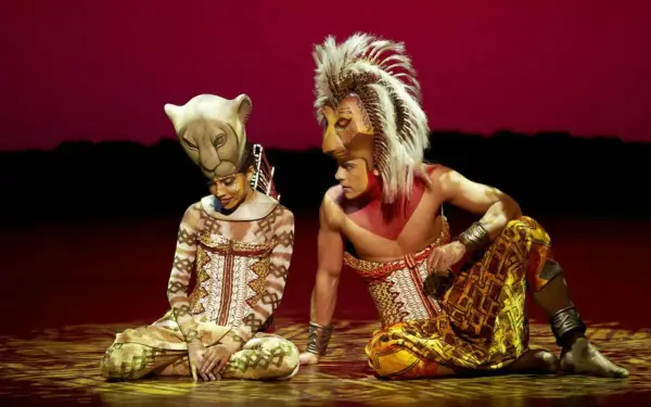 Dos actores con trajes de león, uno con máscara de leona y otro con melena, sentados en el escenario. La duración del Rey León en Madrid es inolvidable.